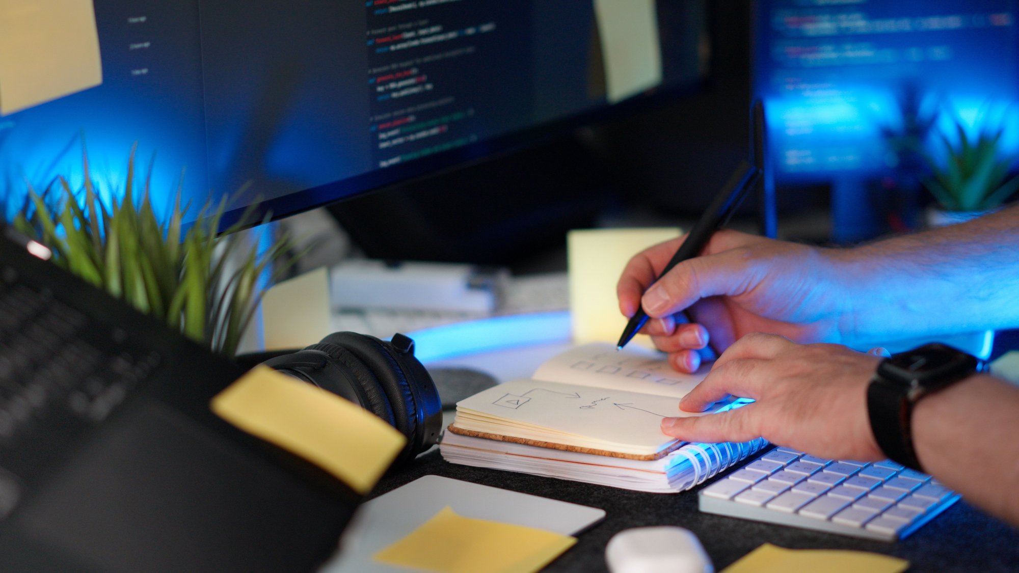 A developer working at a modern desk with a computer displaying code, surrounded by notes and technical work materials. The image represents software engineering, coding, problem-solving, system design, and building software that can adapt to change.